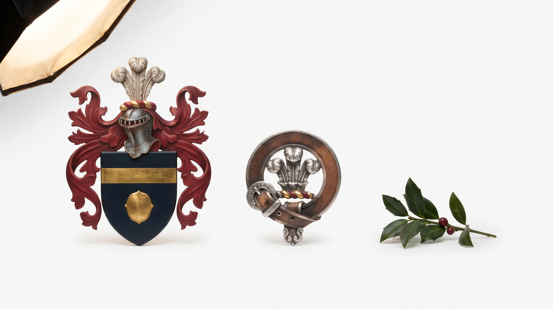 A painted heraldic achievement, a silver crest badge inside a strap and buckle, and a sprig of a plant badge, photographed together at true relative scale