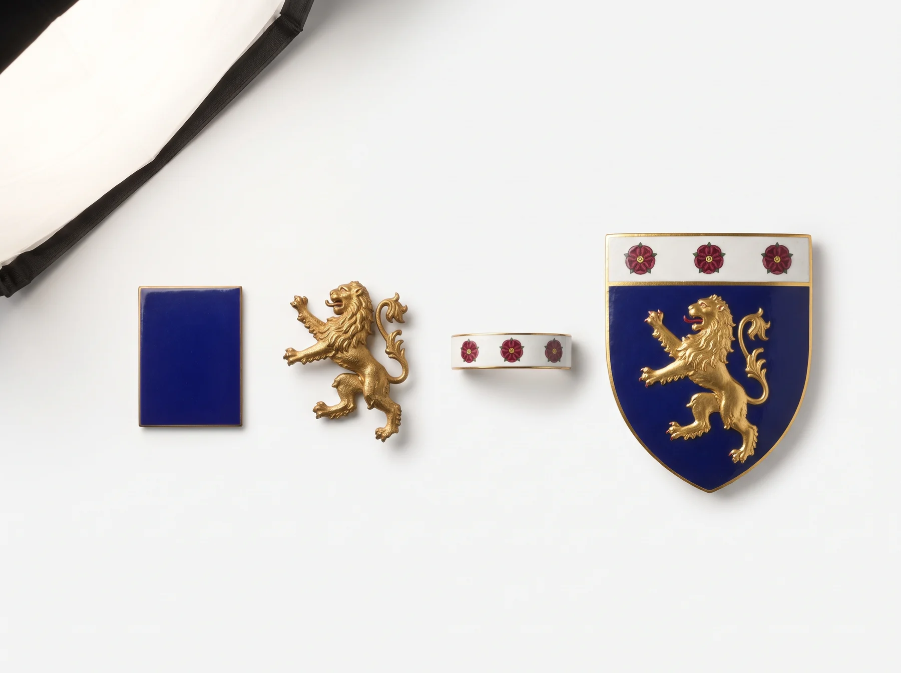 A blue enamel tile, a cast gilt lion and a white band with three red roses laid in a row beside the finished shield that combines all three